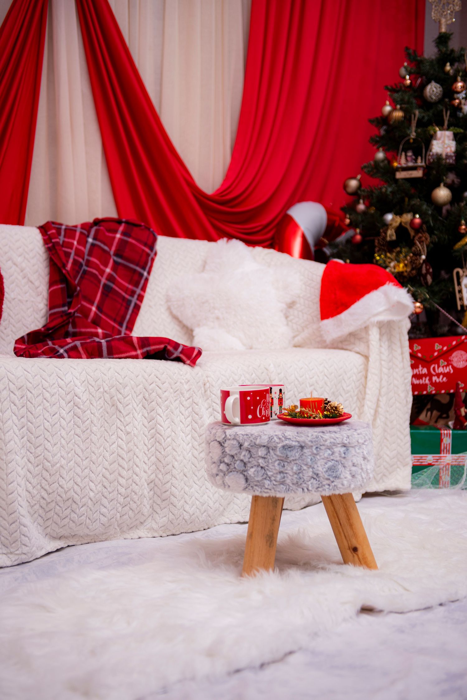 Festive Christmas studio setup with white couch, red curtains, and snowy floor against decorated tree background.