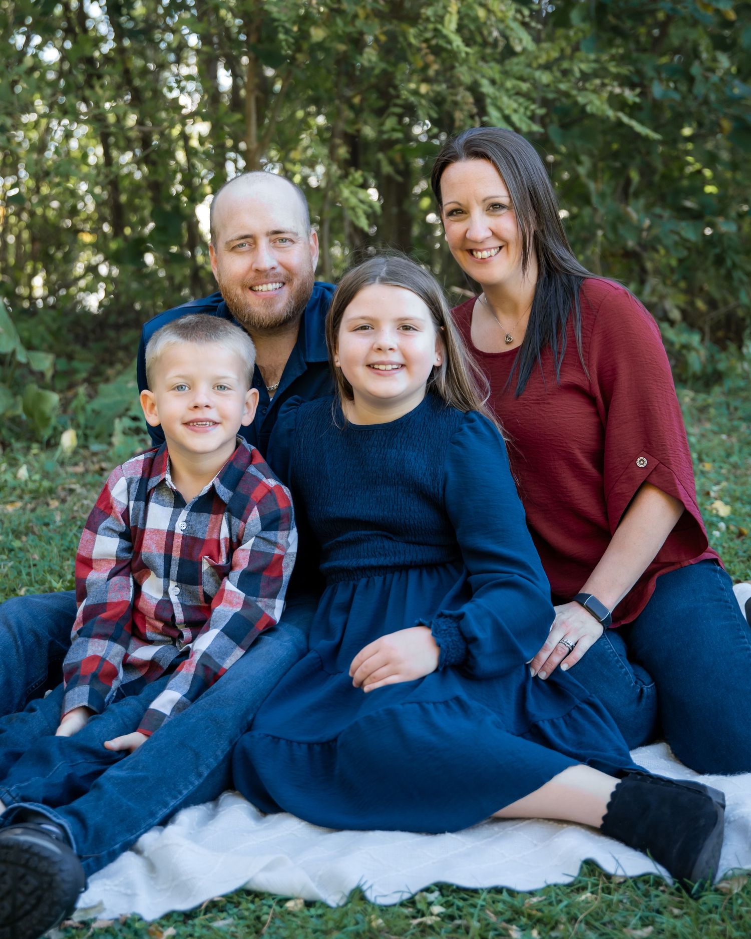 A family sits together outdoors on a white blanket wearing coordinated navy and burgundy outfits in a natural setting.