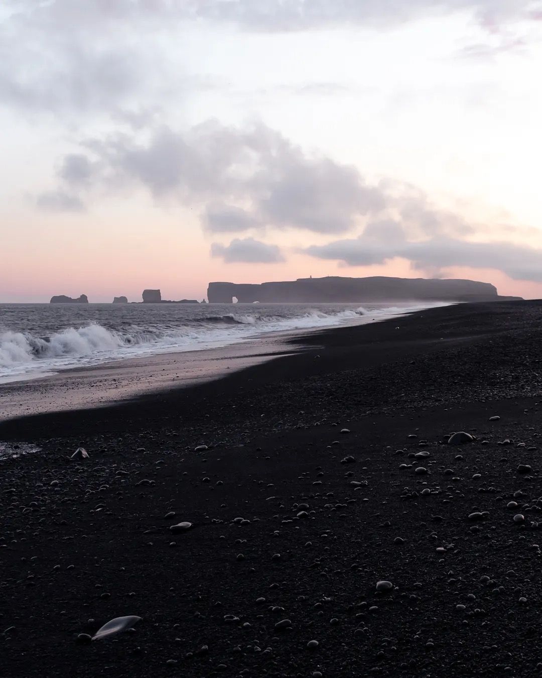 Moody black sand beach with sea mist and distant cliffs at sunrise.
