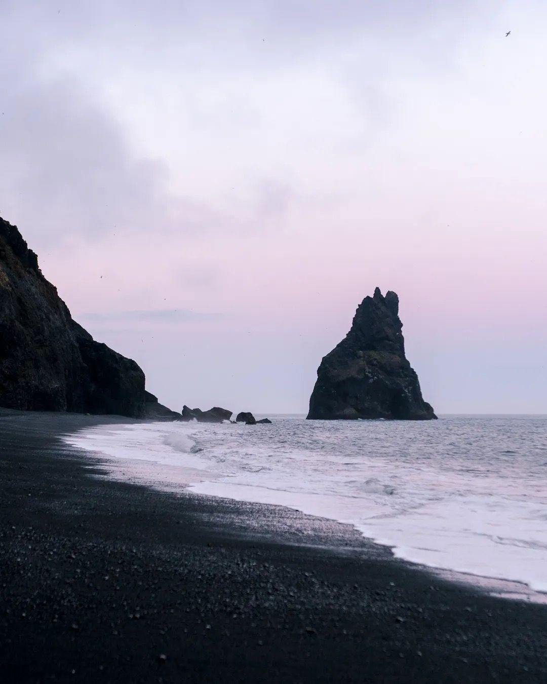 Waves washing onto a black sand beach at sunset with a tall sea stack offshore.