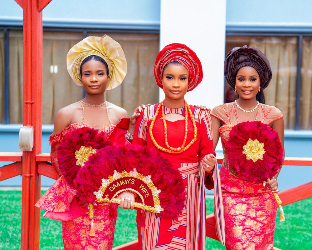 Bridesmaids in vibrant red and gold traditional Nigerian attire stand in line at an outdoor celebration.