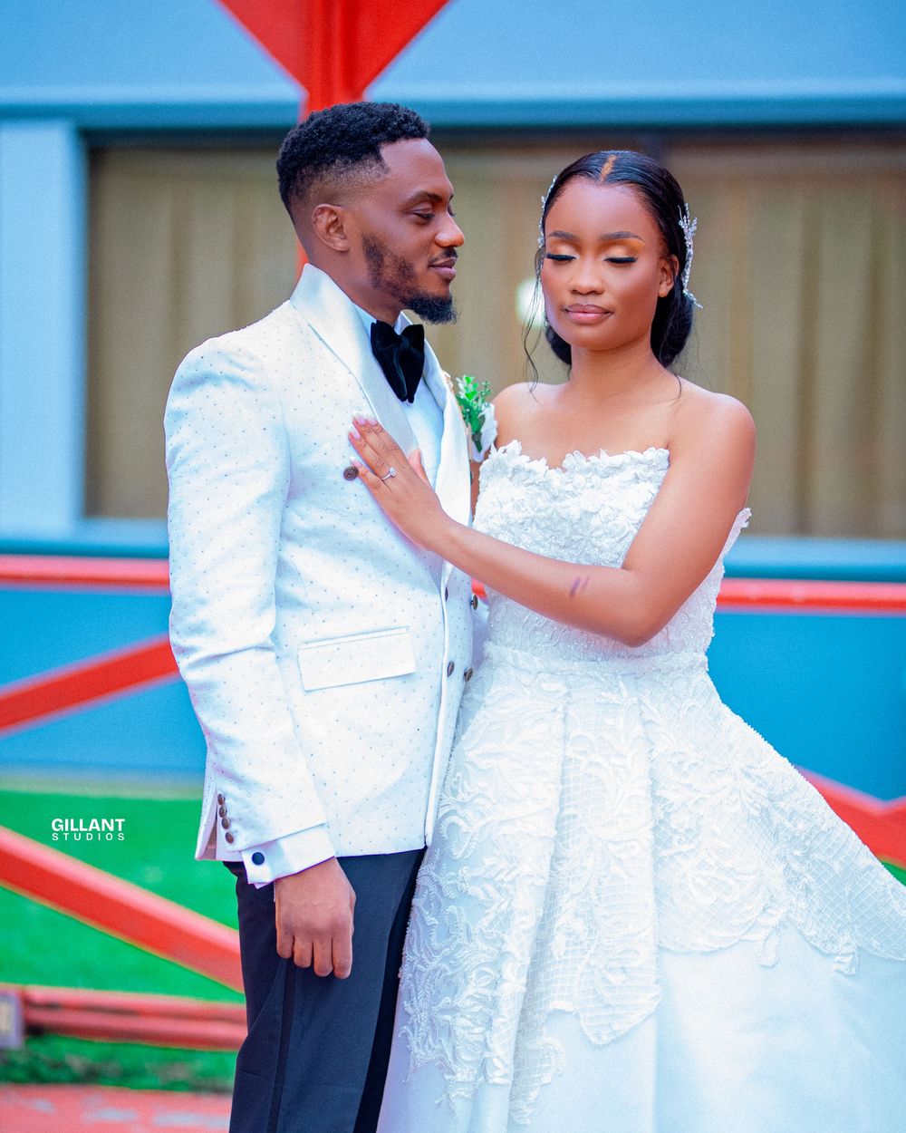A couple in white formal attire shares romantic moments during a photoshoot near blue and red architectural elements.