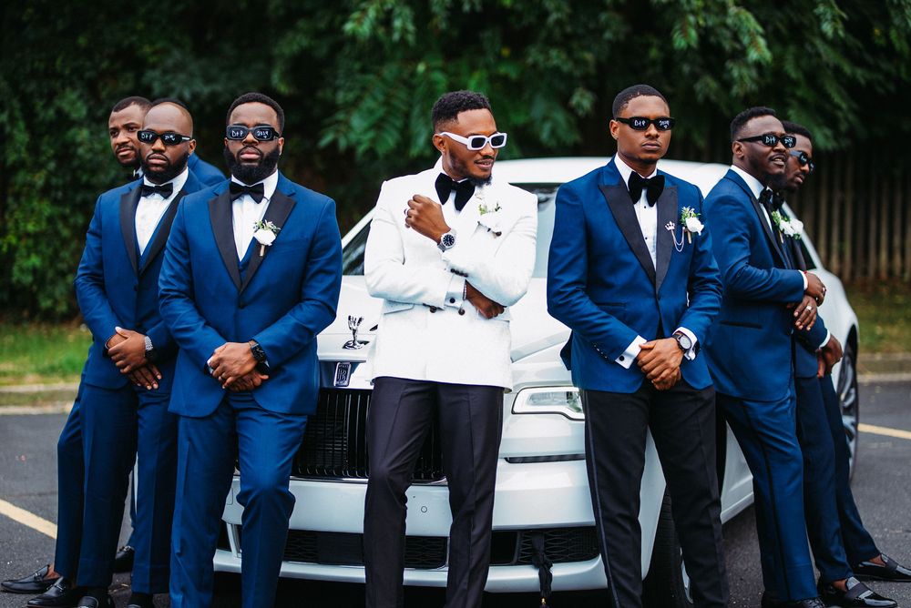 A wedding party group in coordinated blue suits and one white tuxedo jacket pose together in front of a car.