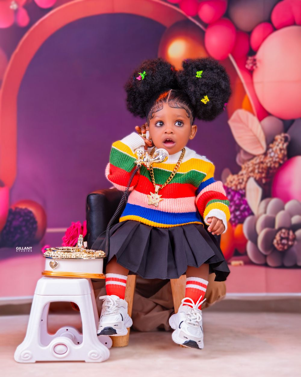 Colorful birthday setup with balloons and teddy bears featuring a toddler in a striped rainbow sweater and black skirt.