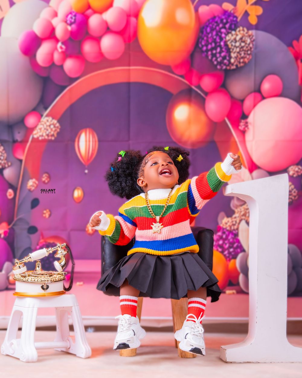 A cheerful child in a colorful striped sweater and skirt poses energetically at a first birthday party with pink balloon decor.