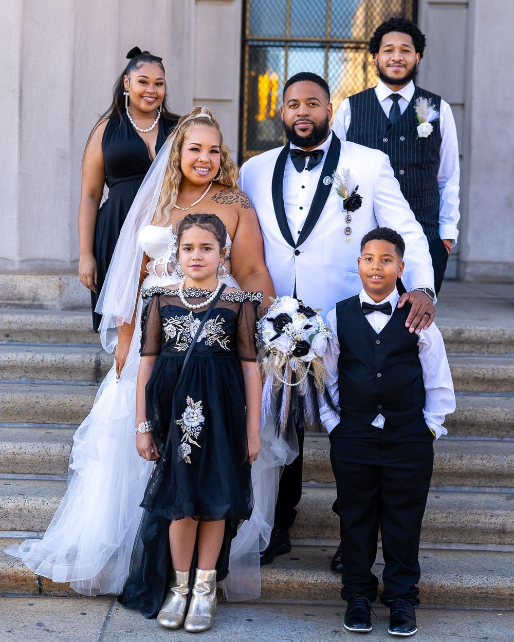 Wedding party stands on stone steps with bride in white gown, attendants in formal attire, and small dog in center.