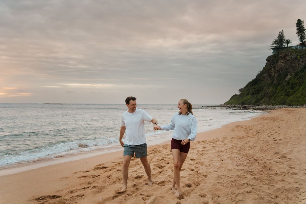 A couple walks hand in hand along a scenic beach at sunrise with waves crashing against the shore at Newport Beach Photoshoot in Sydney