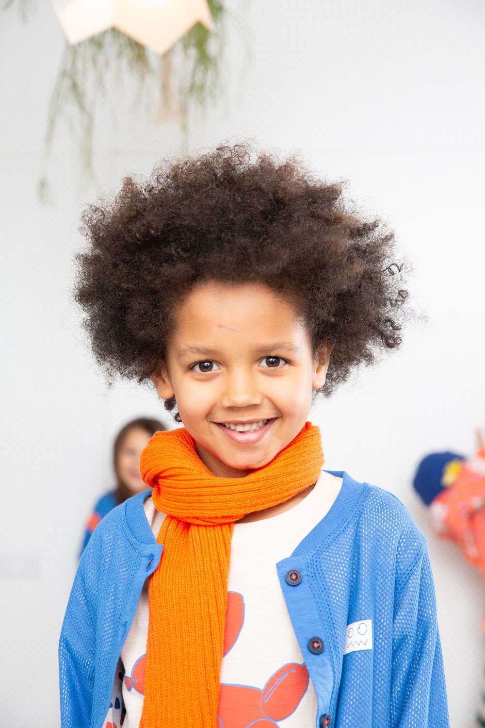 Young child wearing bright orange scarf and blue cardigan against white background.