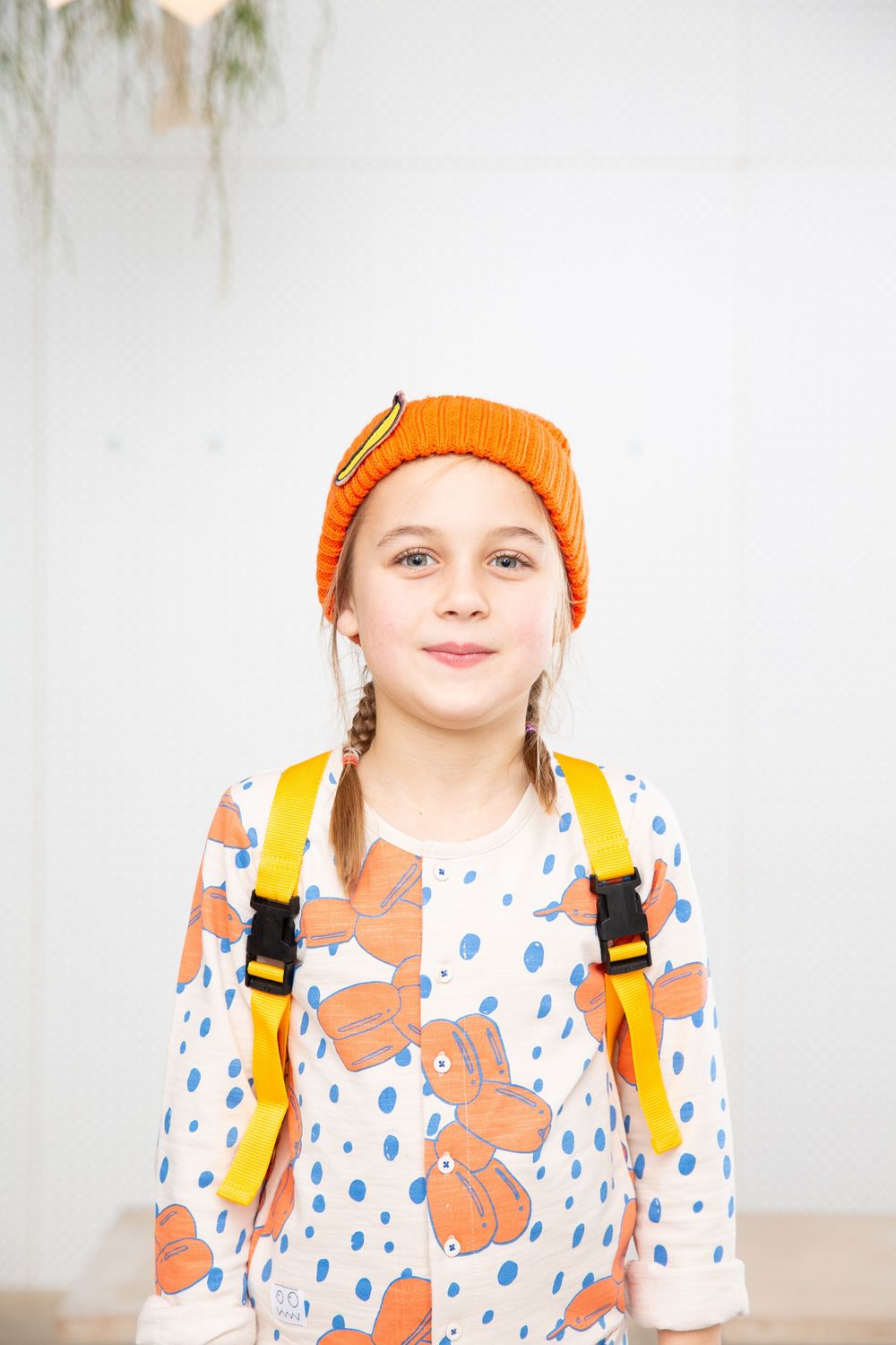 Child wearing orange beanie and polka dot shirt with yellow backpack against white background.
