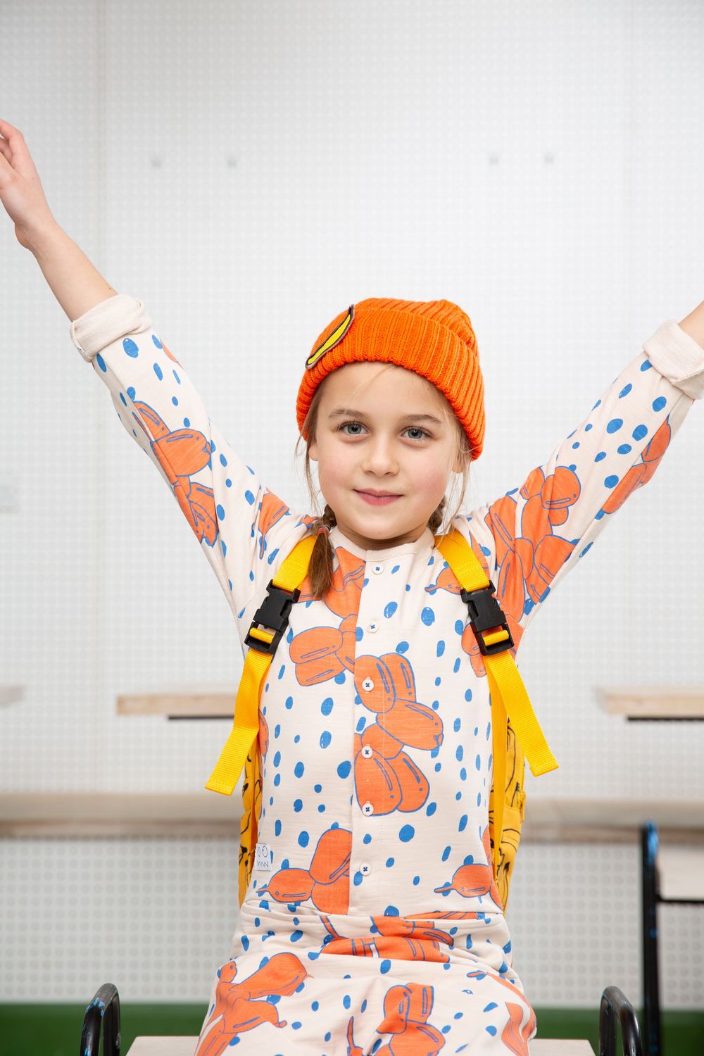 Child in orange beanie and patterned shirt raising arms joyfully against white background.