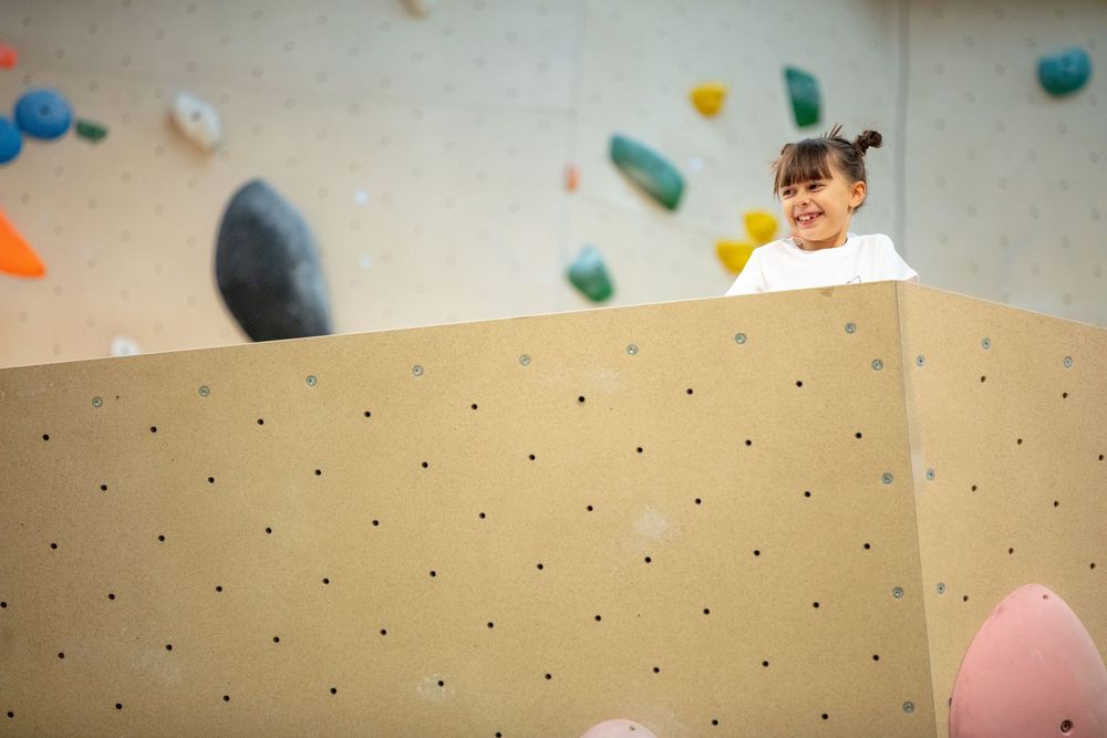Beige indoor climbing wall with scattered colorful holds and safety padding.