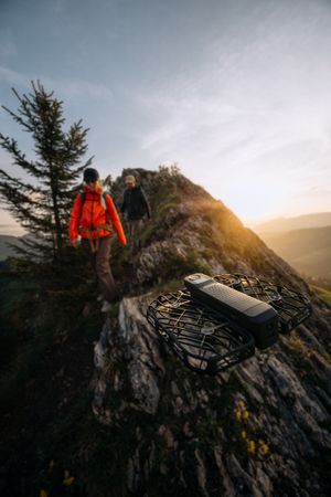 A hiker in bright orange jacket stands on a rocky mountain peak during golden sunset hour while exploring wilderness terrain.