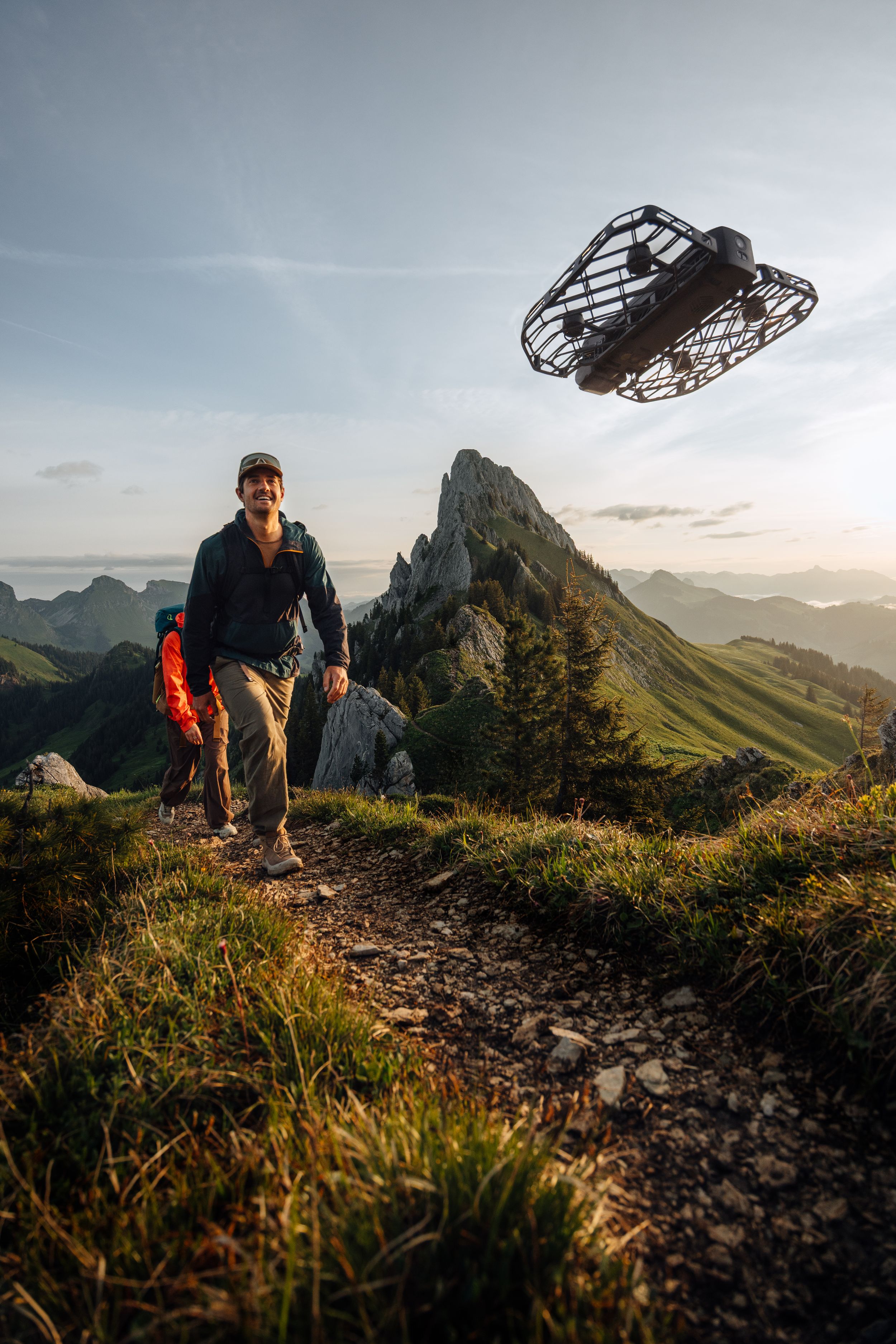 Hikers traverse a scenic mountain trail at sunset with dramatic peaks and valleys stretching into the distance.
