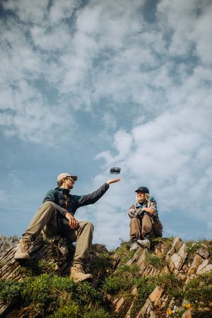 Two hikers sharing a drink while resting on a rocky hillside under a cloudy blue sky.