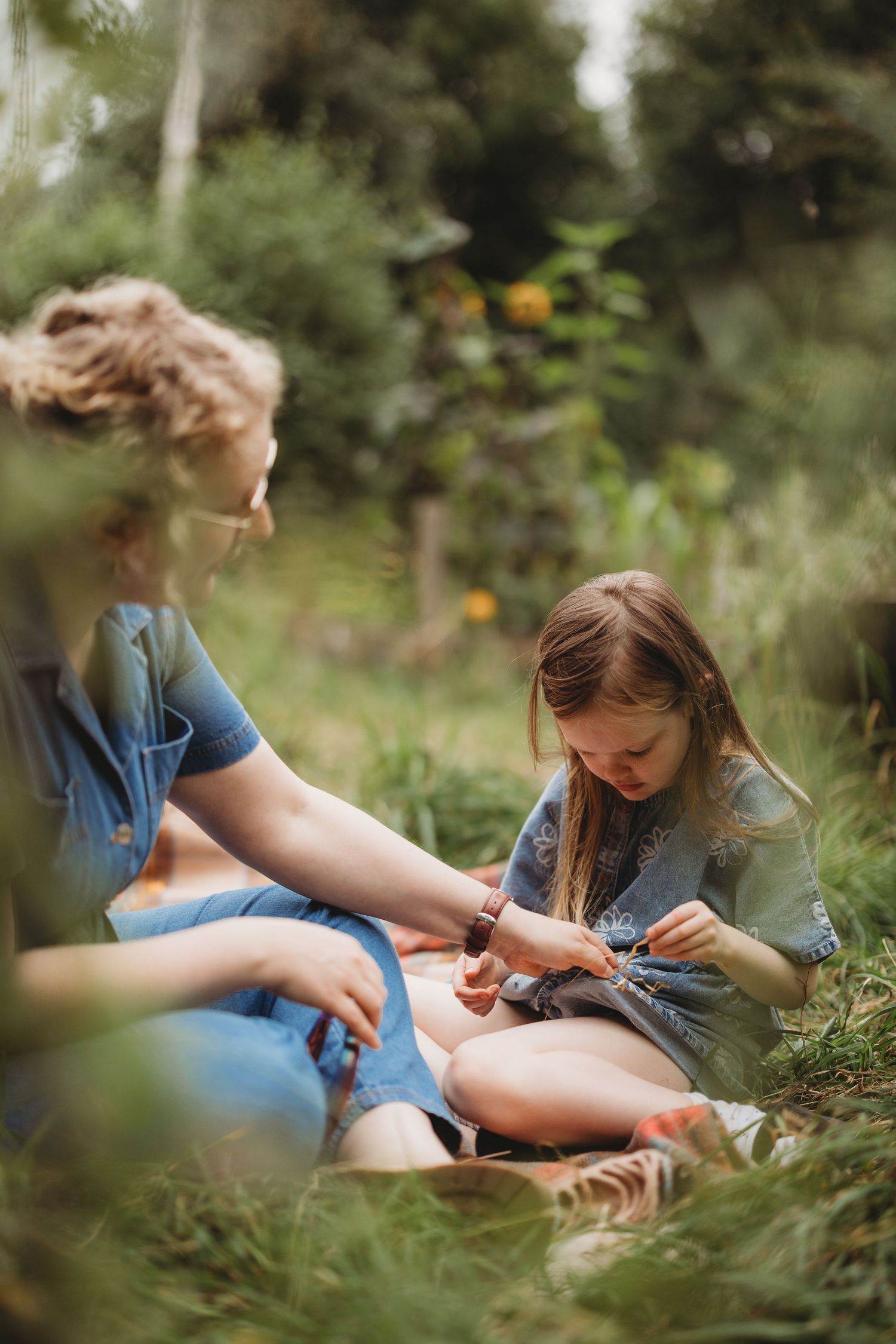 A person sits in a garden meadow wearing denim, gently interacting with plants and flowers in a peaceful outdoor setting.