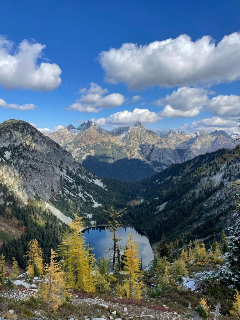 Elopement photographer on Scenic mountain landscape with alpine lake and golden larch trees in autumn against rugged peaks and blue sky.