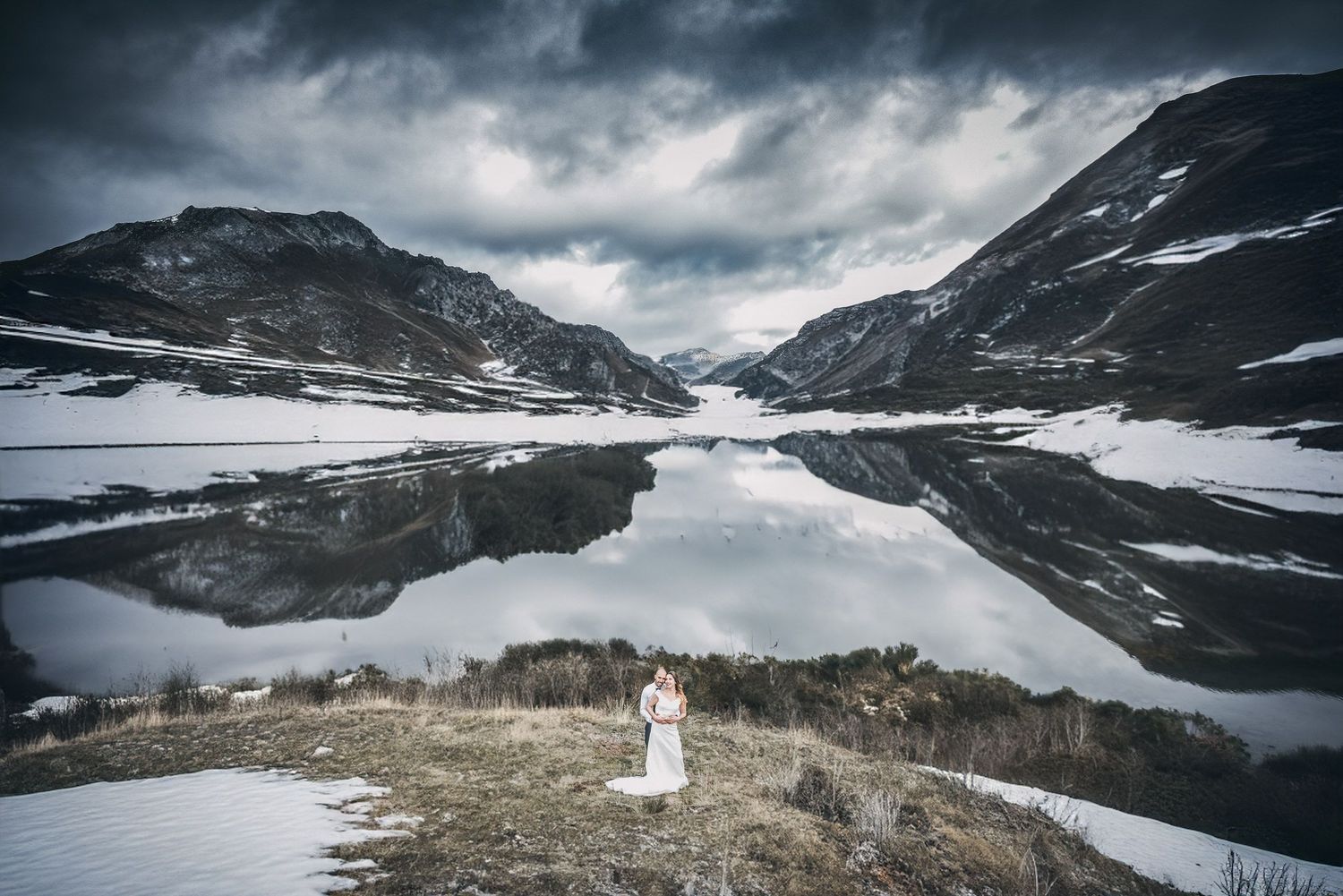 A dramatic mountain landscape series with snow patches and moody clouds showcases a couple in wedding attire exploring nature.