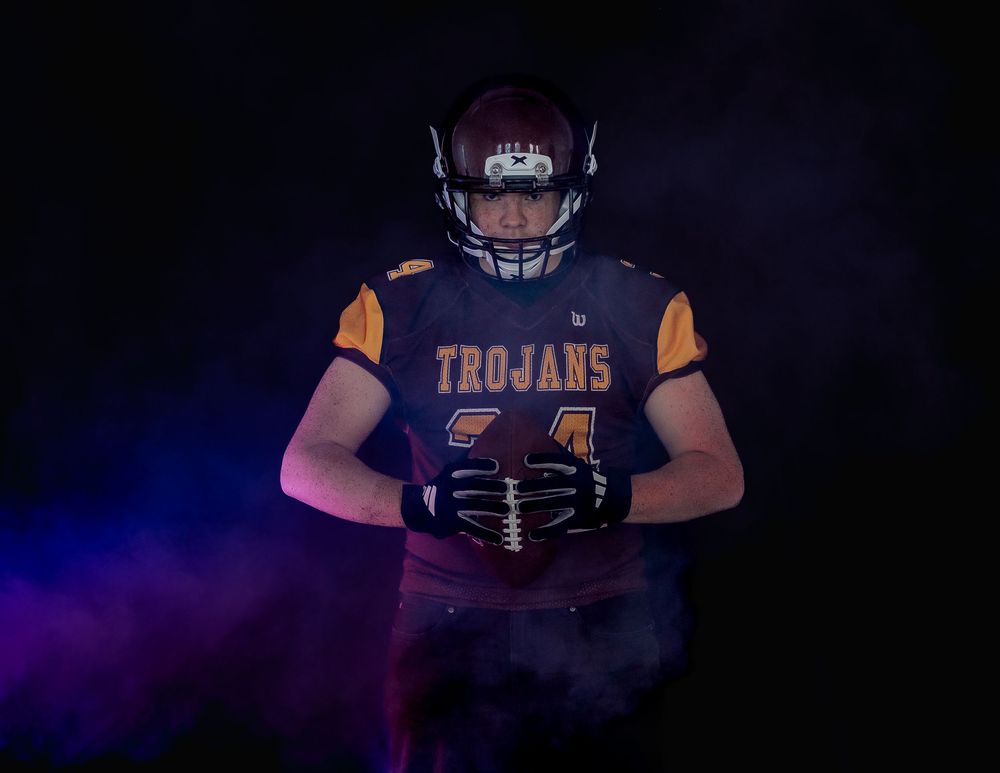 Football player in black and gold uniform stands in dramatic lighting with crossed arms against dark background.