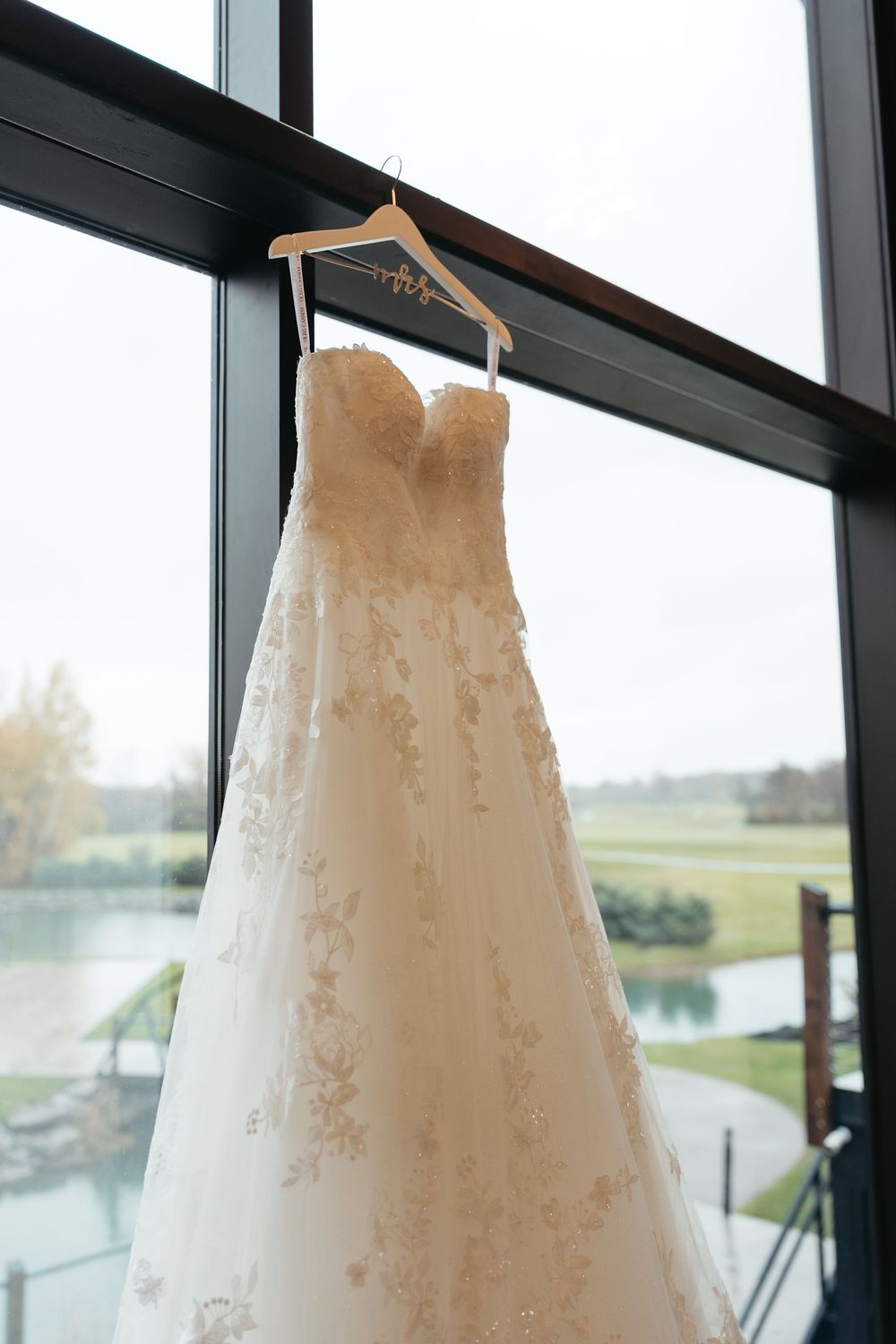 Three elegant white wedding dresses with delicate lace details hang in a row against large windows overlooking a scenic view.