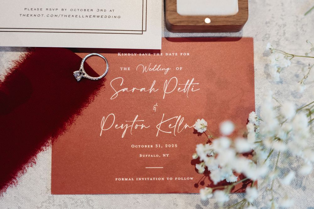 Wedding invitation in burgundy with white calligraphy text and engagement ring styled with delicate white flowers.