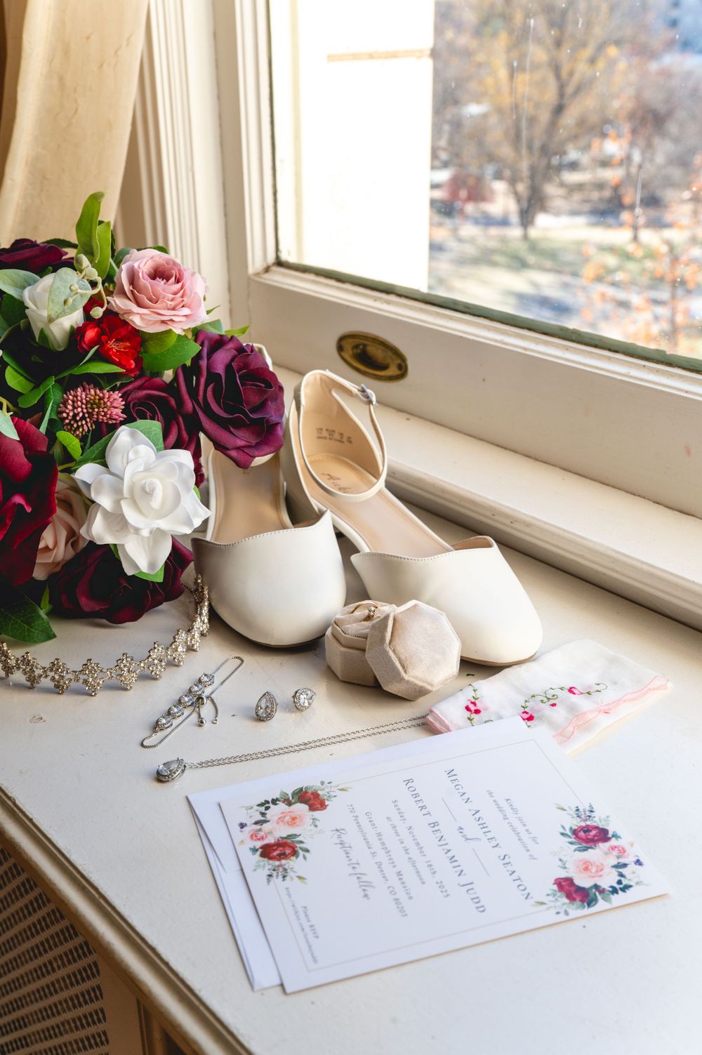 Wedding invitation suite with floral design displayed on windowsill alongside bridal accessories and fresh flowers.