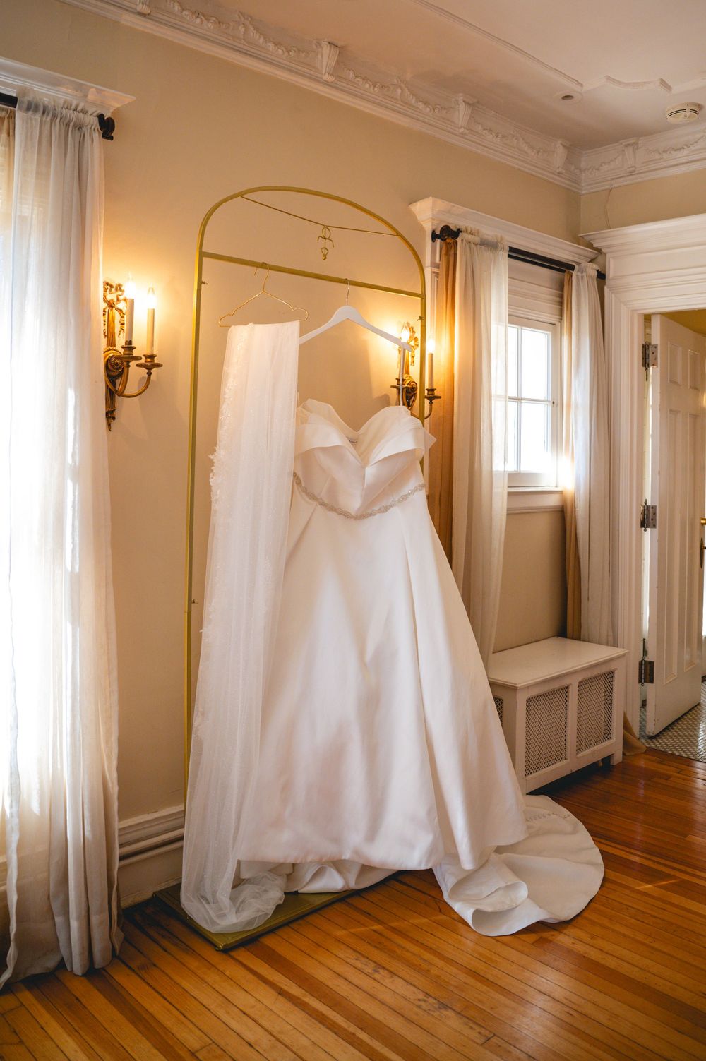 An elegant white wedding dress hangs on a gold arch frame in a room with ornate wall sconces and hardwood floors.