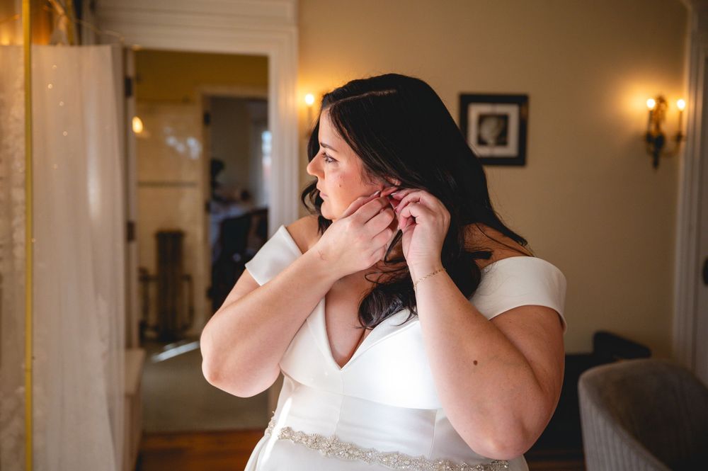A bride in a white wedding dress gets ready in a warmly lit room with elegant wall sconces and framed artwork.