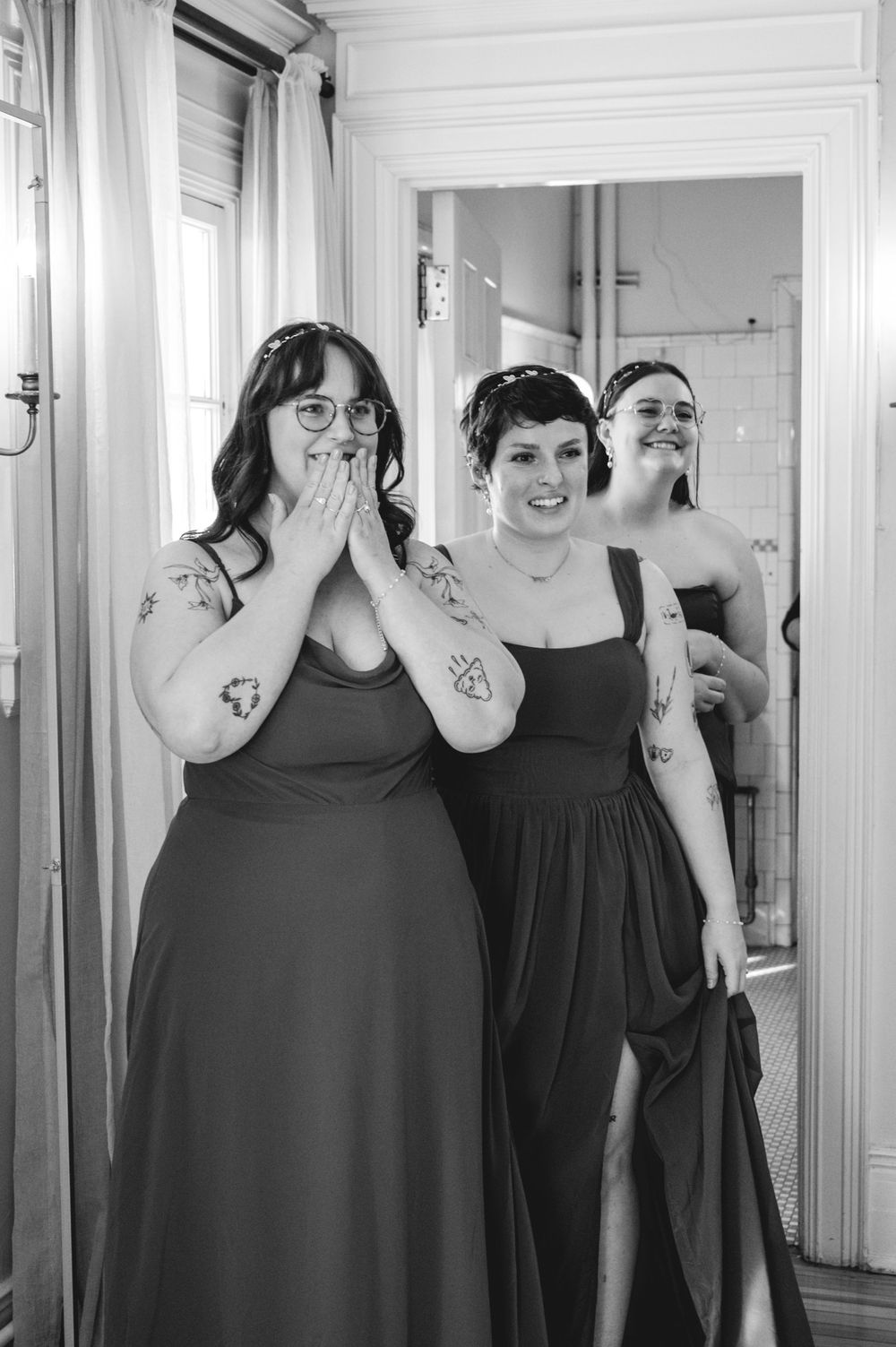 A group of bridesmaids in black dresses cover their faces while posing in a white hallway.
