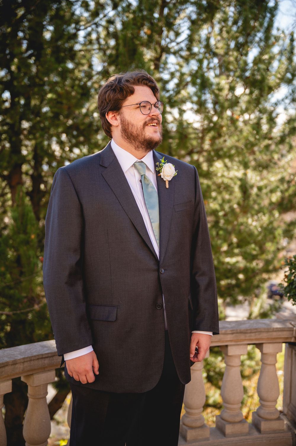 A groom in a dark suit and light blue tie smiles while standing on a balcony with stone railings.