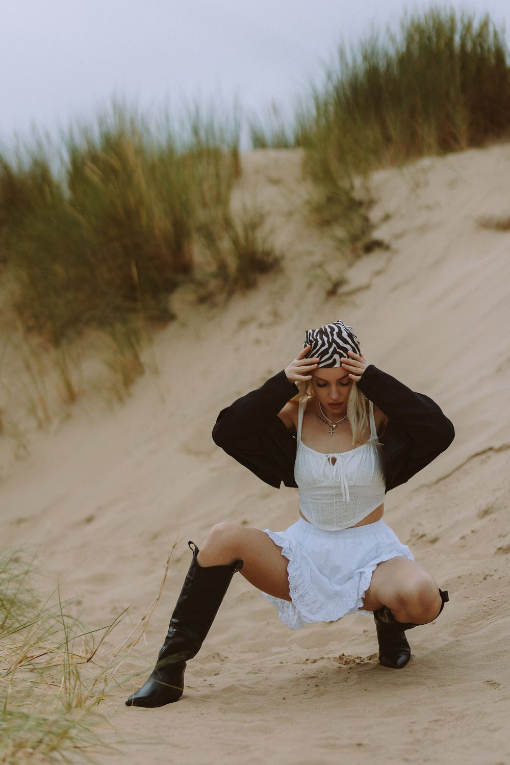 A figure in a black jacket and white top poses dramatically on sandy dunes with beach grass in the background.