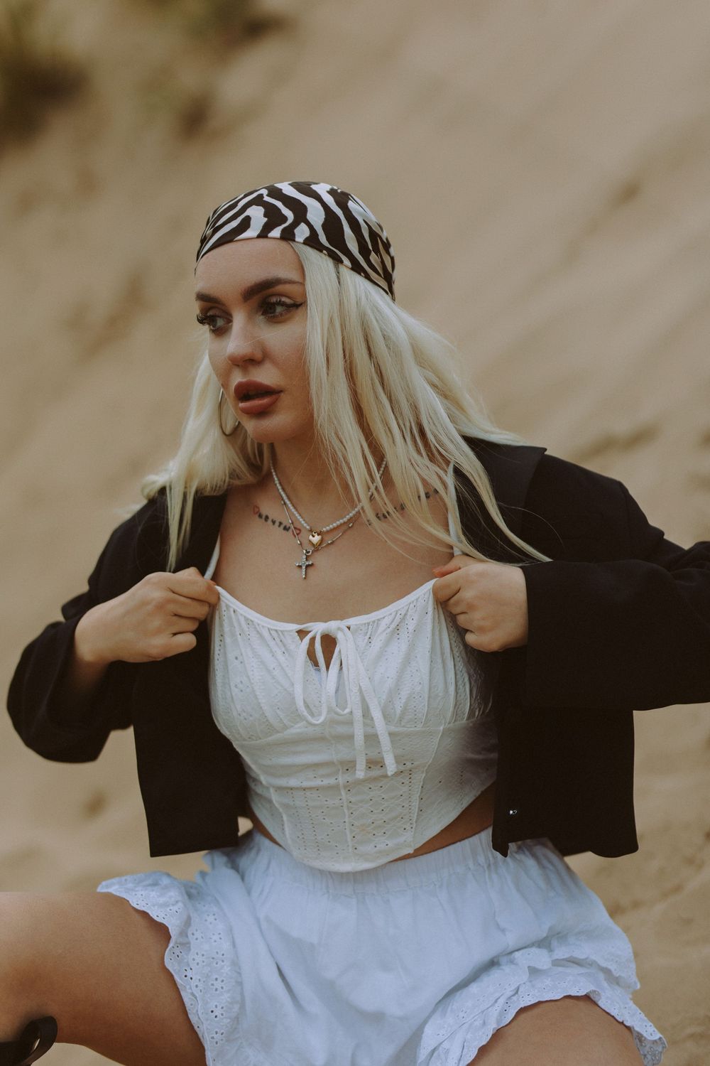 A person in a white corset top and black cardigan poses against a sandy backdrop in a vintage-inspired photoshoot.