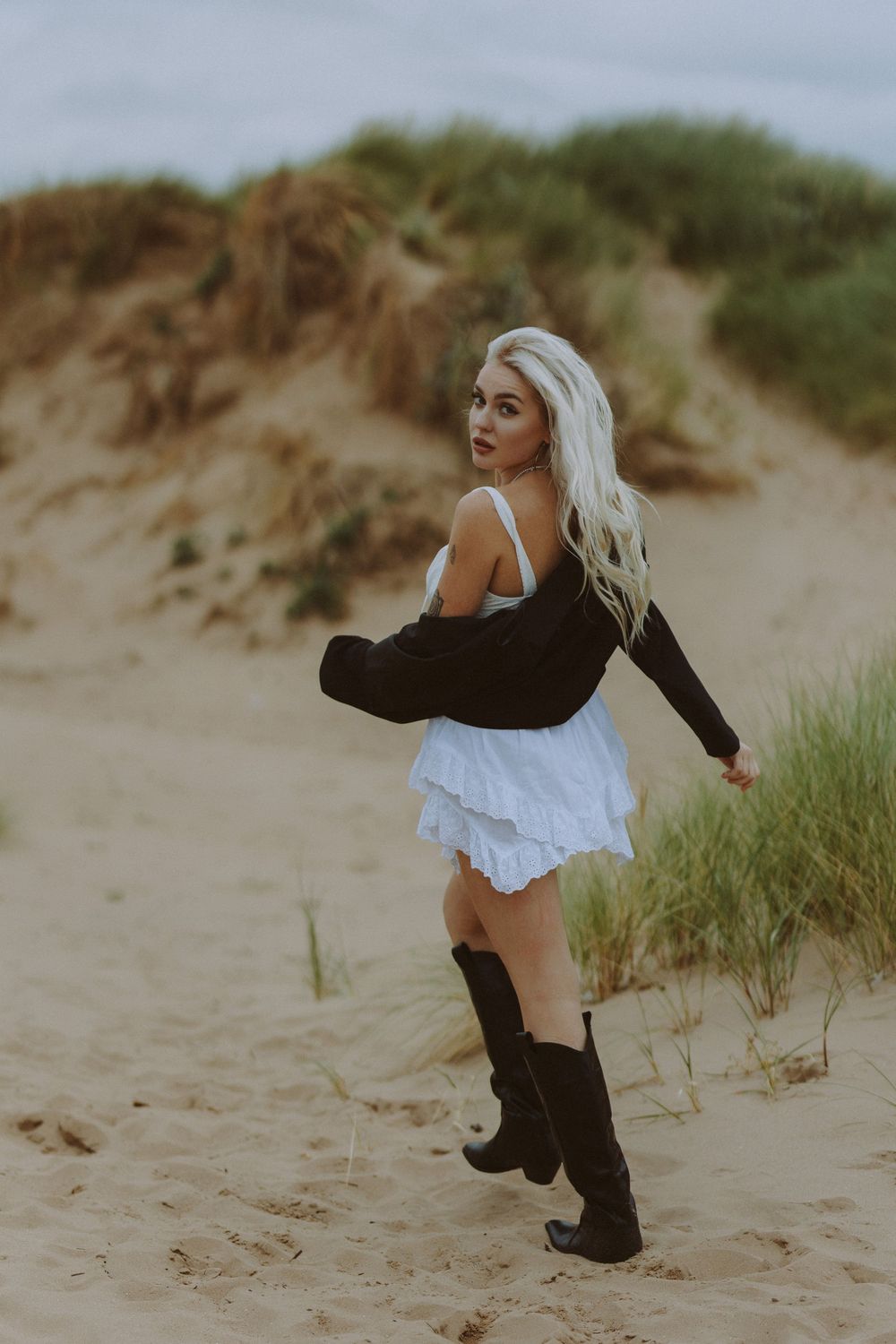 A figure in black boots and white dress poses gracefully against sandy dunes and beach grass at sunset.