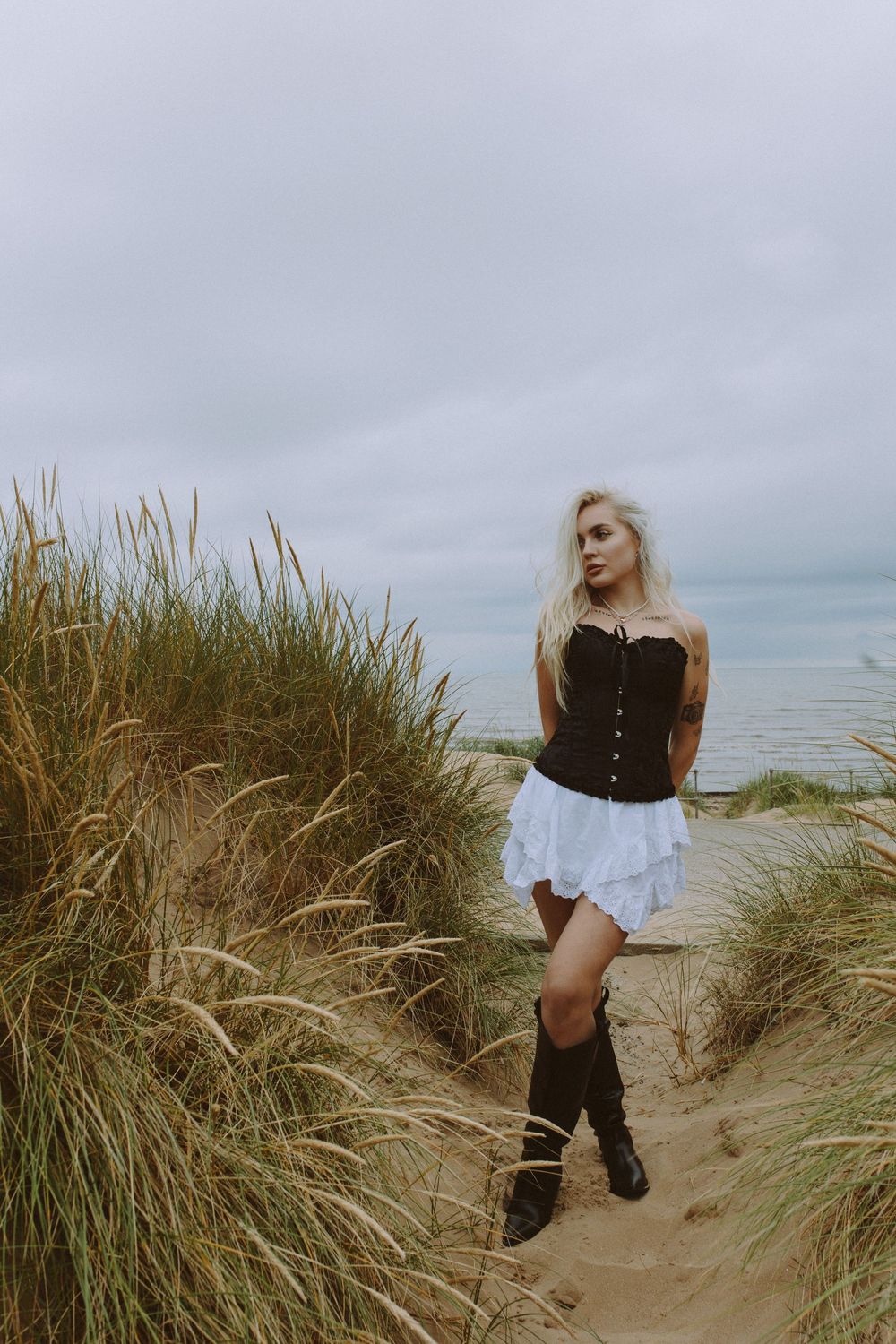 A series of five photos showing a person in dark clothing posing among tall beach grass and sand dunes on an overcast day.