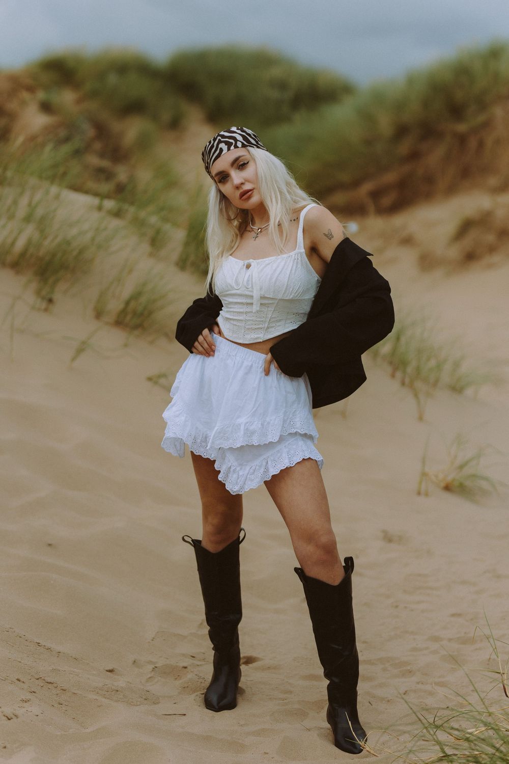 A person in a white dress and black boots poses dynamically on sandy beach dunes with coastal grass in the background.