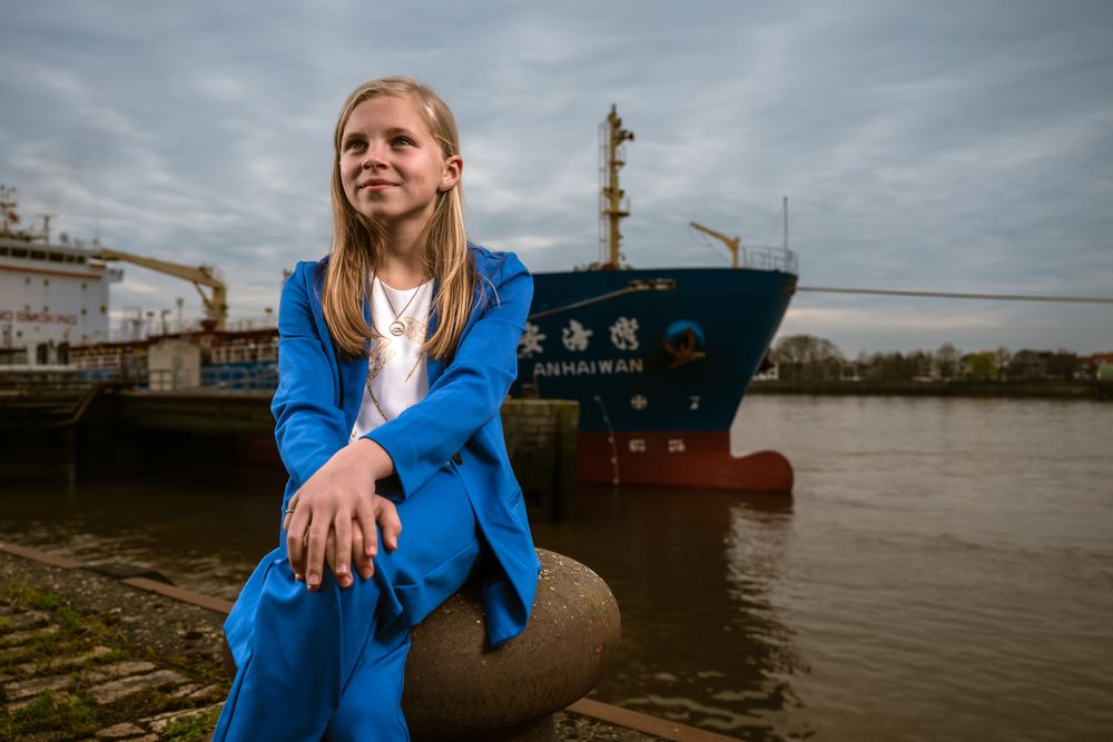 Meisje in blauw kostuum poseert zelfverzekerd aan de Schelde tijdens communieshoot – buitenlocatie door Bart Boodts Photography