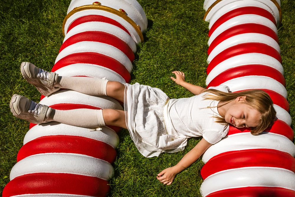 Meisje in witte jurk lacht liggend op rood-witte vormen tijdens een vrolijke communie fotosessie – creatieve fotografie door Bart Boodts