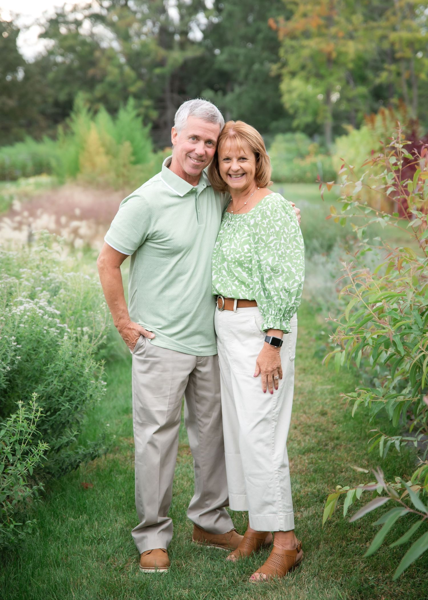 A couple in coordinating green and white outfits poses together in a lush garden setting with wildflowers.
