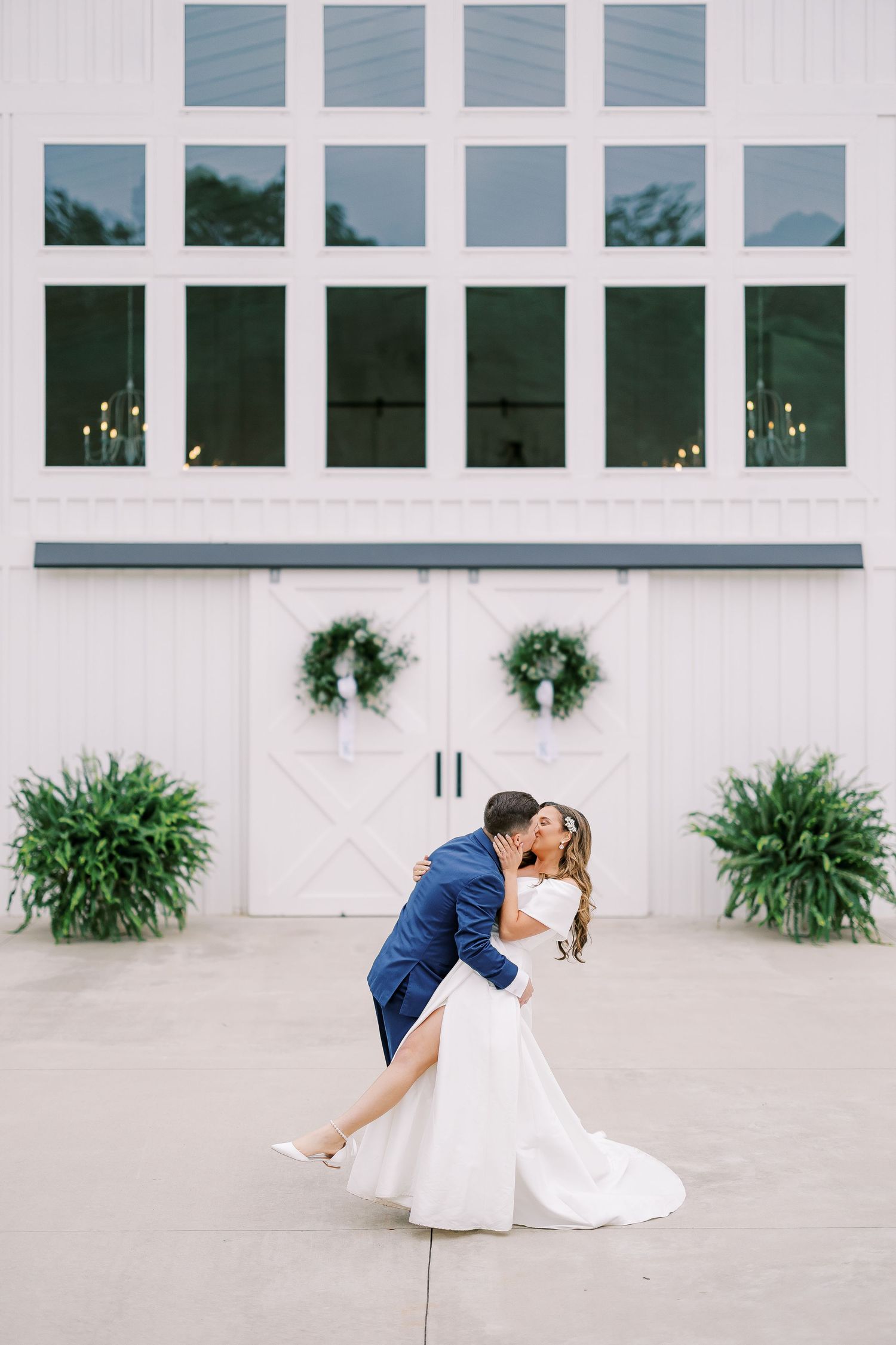 A couple in wedding attire share a romantic dance dip pose outside a modern white building with large windows.