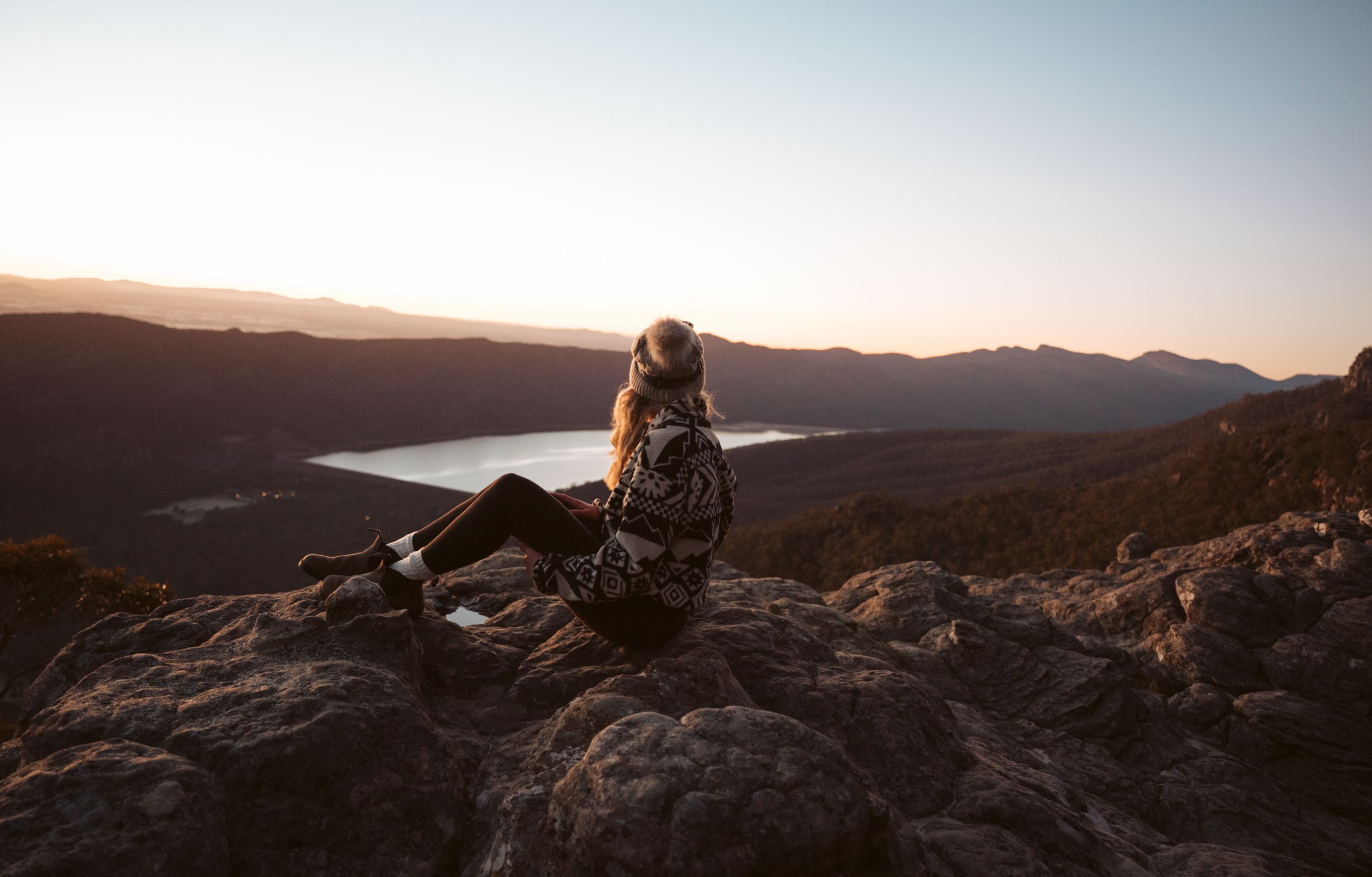 Woman sitting on rocky outcrop watching sunset over lake and mountain ranges in Grampians Victoria