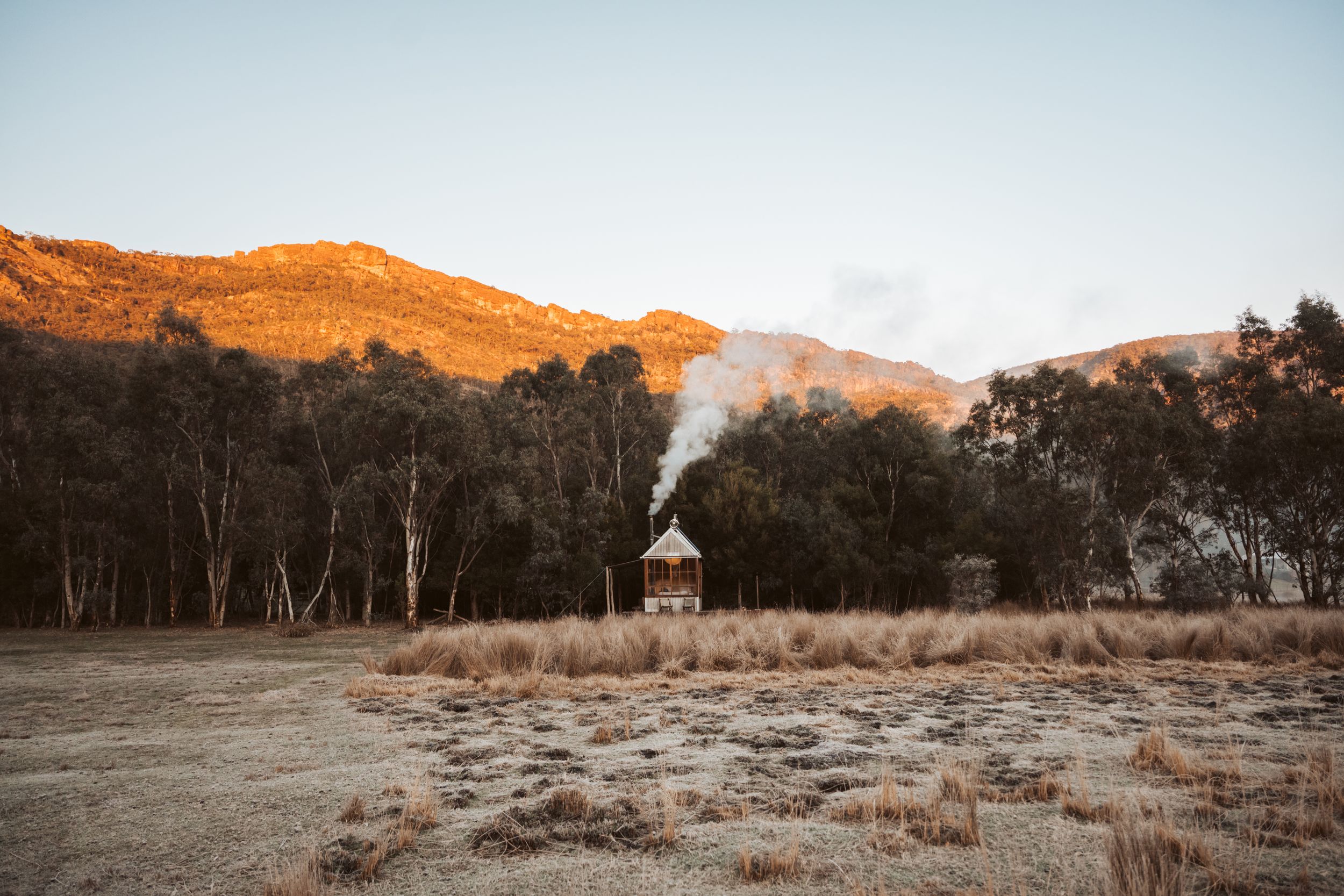 Remote wooden cabin with chimney smoke beneath golden mountain ridges at sunrise in Grampians Victoria