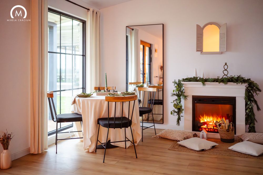 Cozy dining room with round table, wooden chairs, and glowing fireplace decorated with greenery and floor cushions.