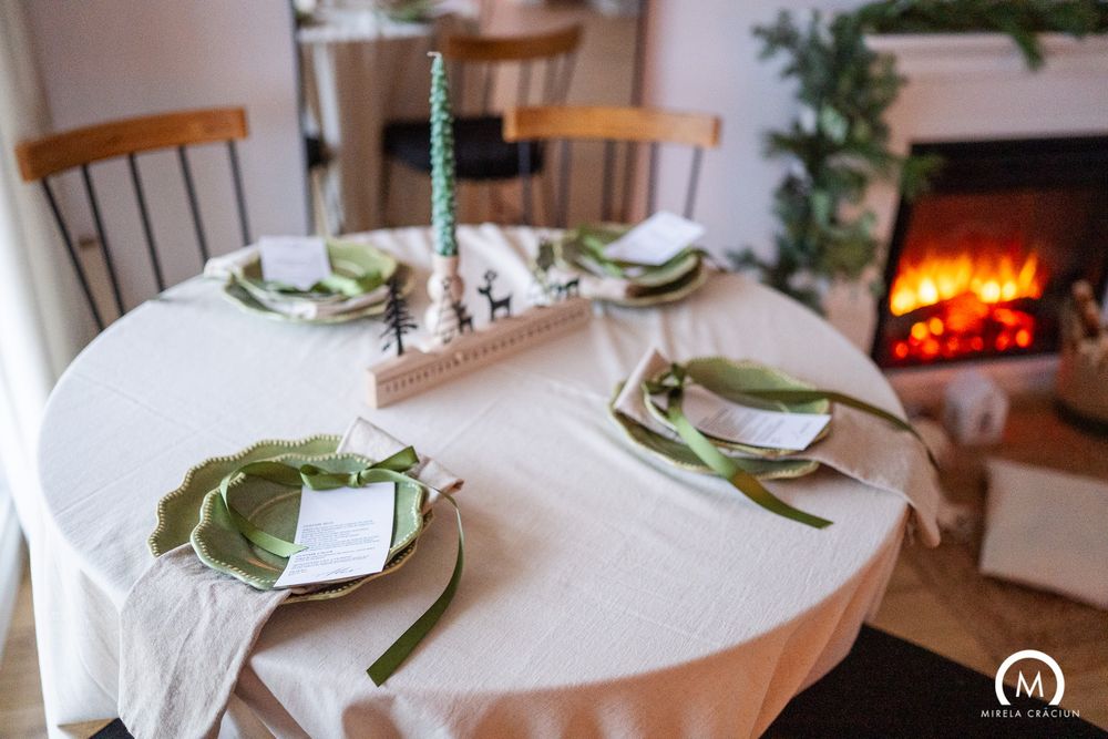 Elegant table setting with white tablecloth, green napkins and plates near a warm fireplace with festive decorations.