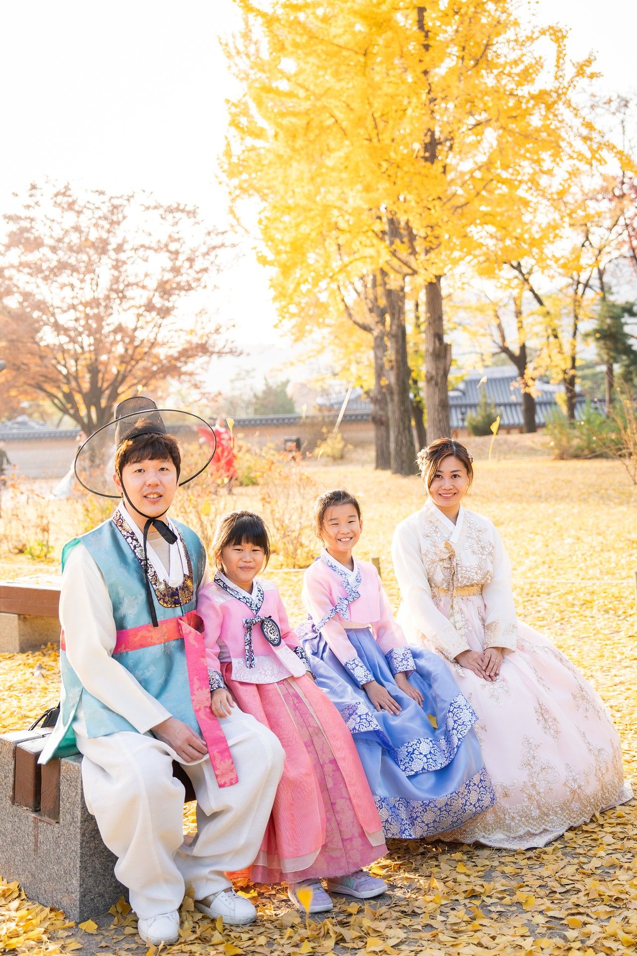 A group in traditional Korean hanbok attire sits on stone benches surrounded by golden autumn leaves.