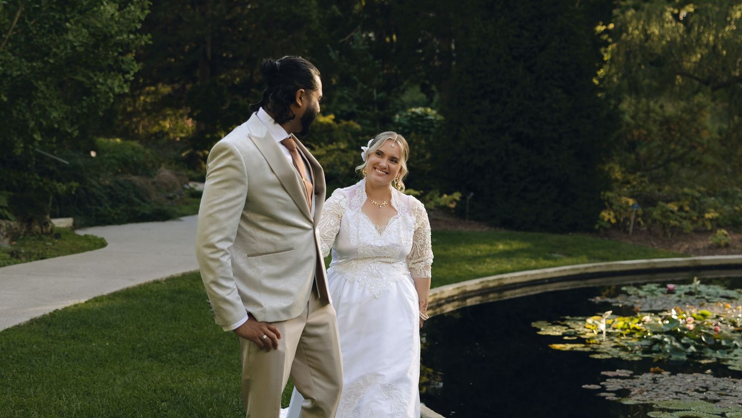 A wedding couple poses outdoors in front of a scenic garden pond with lush greenery and landscaping.