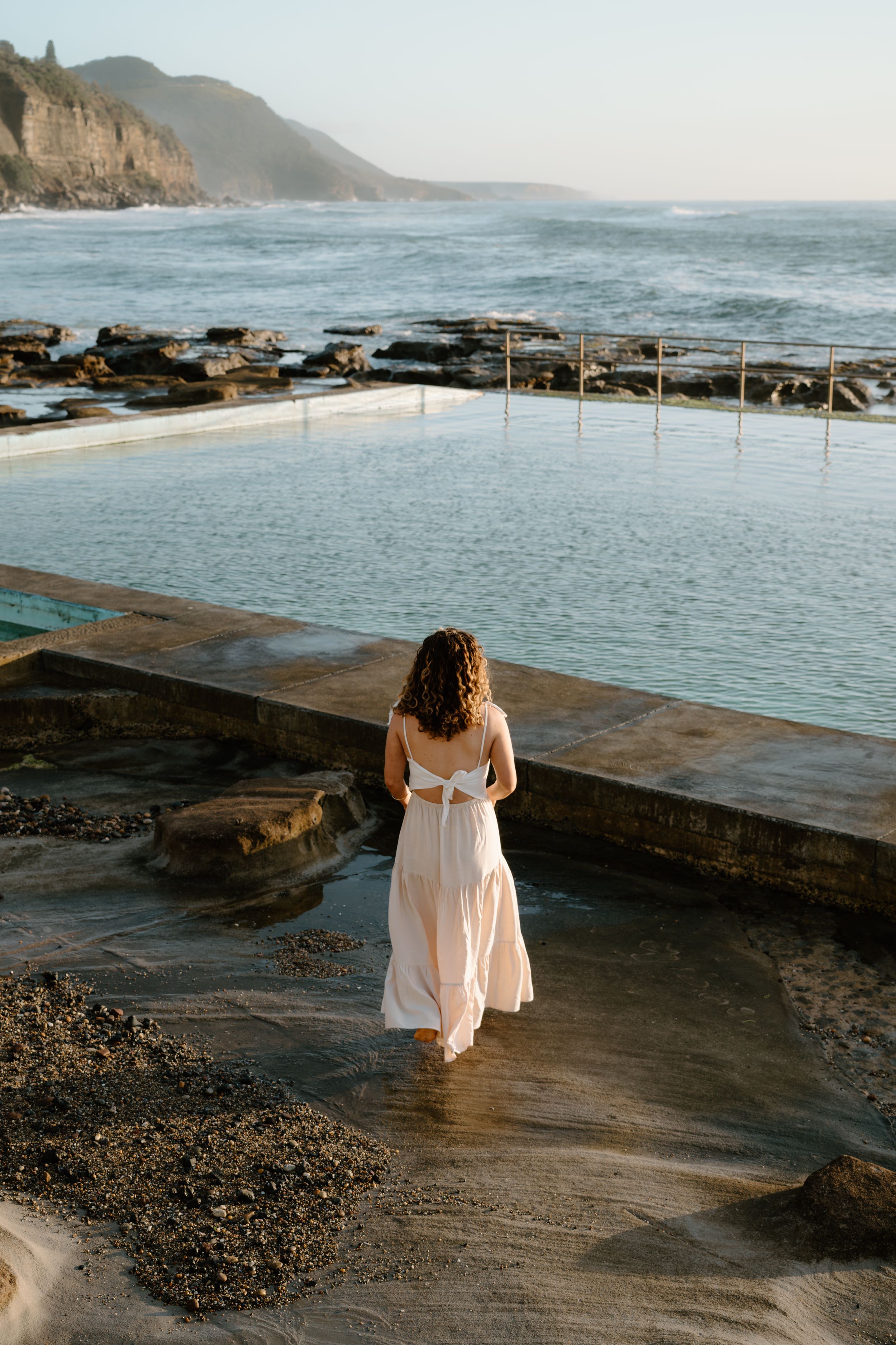A scenic ocean pool and rocky coastline at sunset with a white dress visible in the foreground.