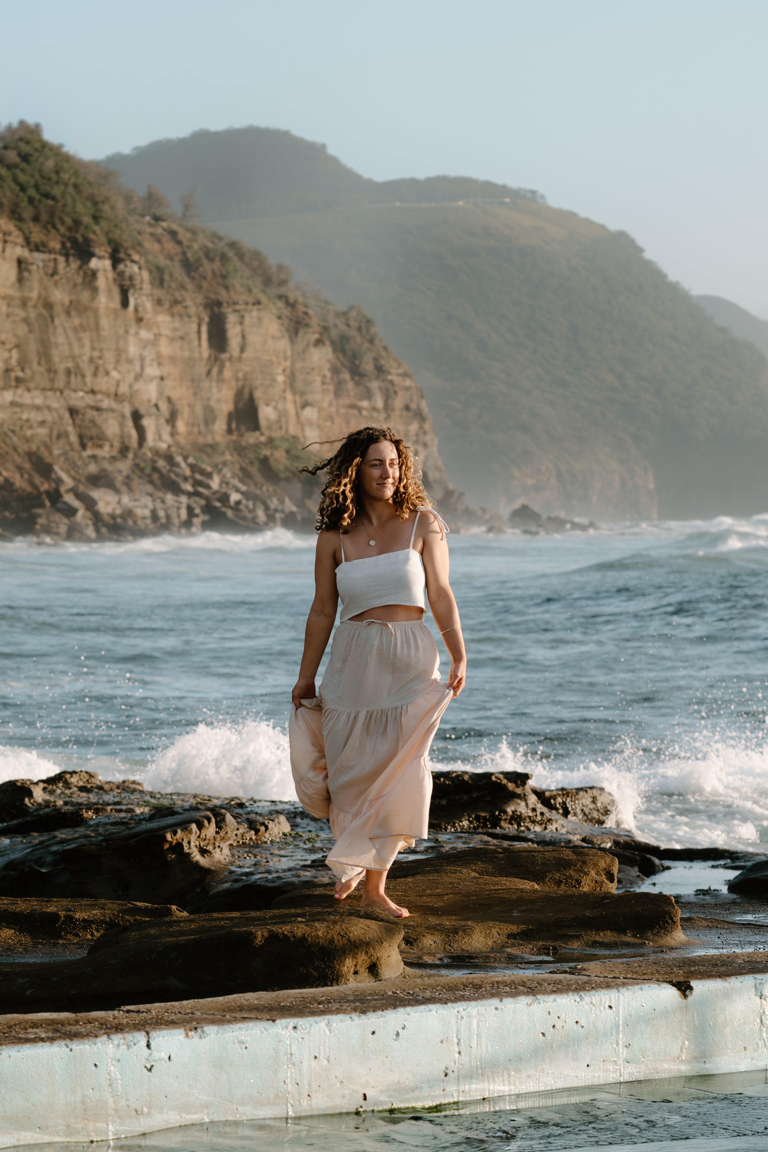 A person in a flowing white dress poses gracefully on rocky coastal cliffs as ocean waves crash against the shore.