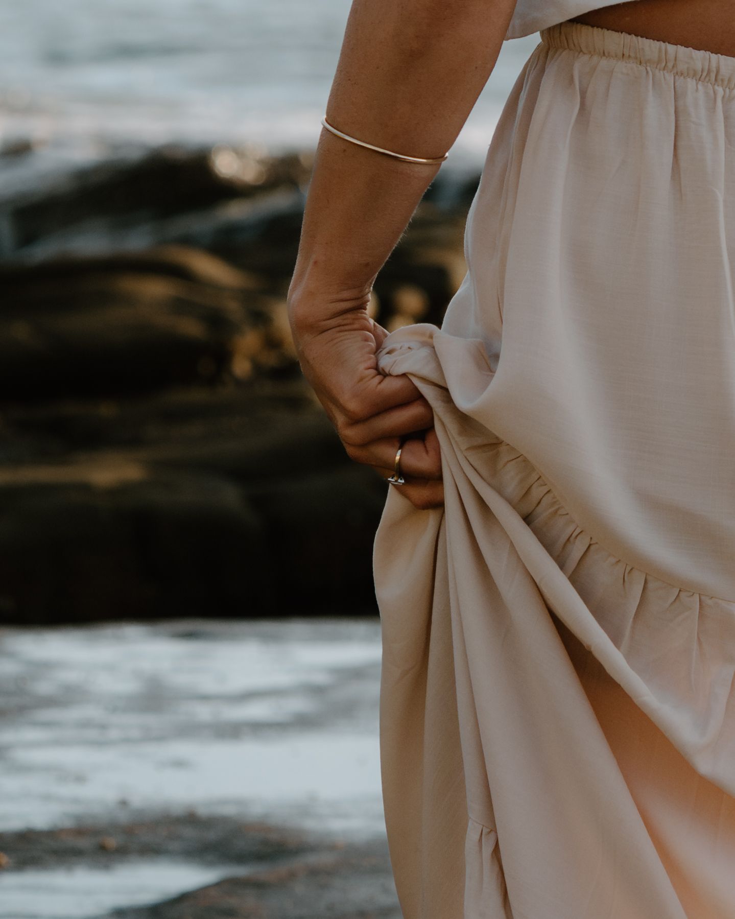 A delicate cream-colored dress flows in the breeze against a rocky coastal backdrop at sunset.