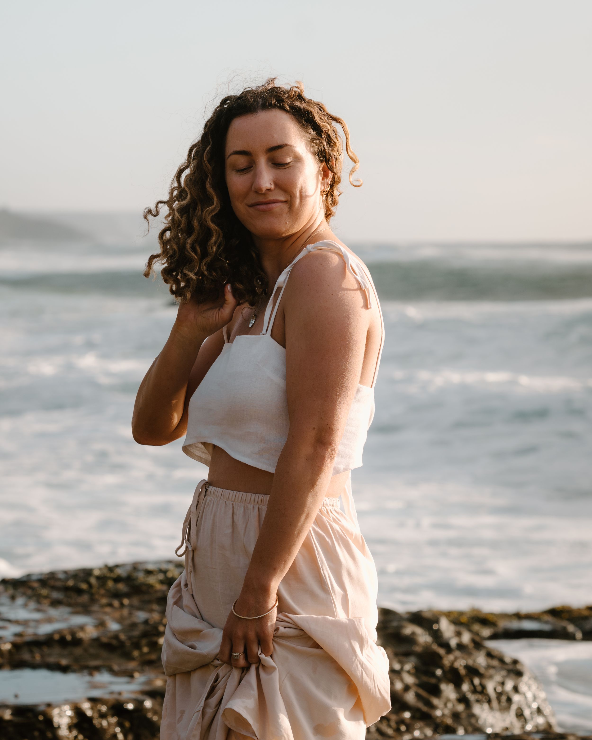 A woman in a white dress poses thoughtfully against a rocky coastal backdrop during golden hour.