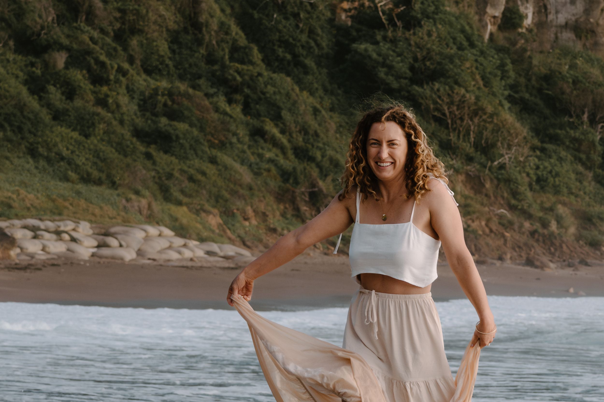 Woman in white crop top and flowing skirt twirls on a scenic beach with cliffs and waves in the background.