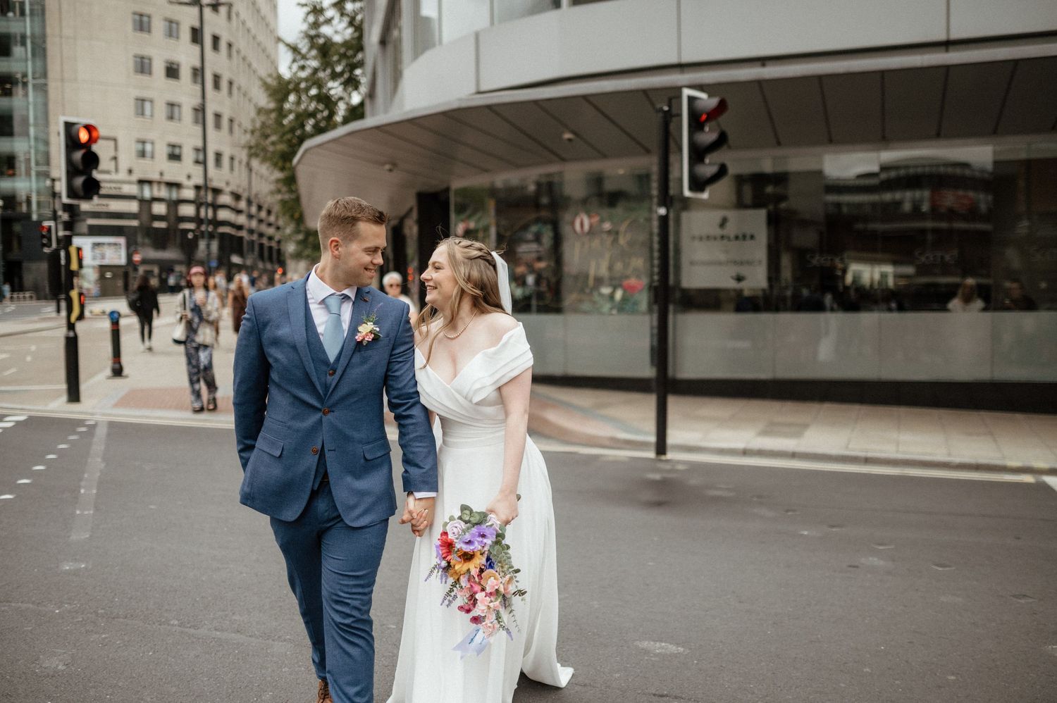 A couple in wedding attire walks together on an urban street with tall buildings in the background.